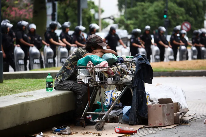 An unhoused man sits outside Argentina’s Ministry of Human Capital during a protest against food scarcity. He carries his belongings in a shopping cart, leaning his arms against it as he watches the proceedings. Behind him, riot police stand in a line, holding their riot shields in front of them.